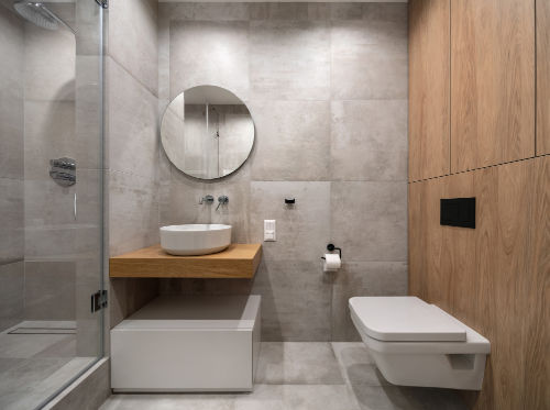 Luminous modern bathroom with textured beige tiled walls and floor. There is a glass shower cabin, wooden rack with a white sink, round mirror, stand, lockers, toilet. Horizontal.